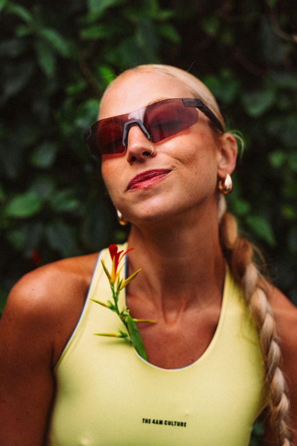 Mulher loira de óculos e regata amarela, com uma flor presa na alça, em frente a folhagens