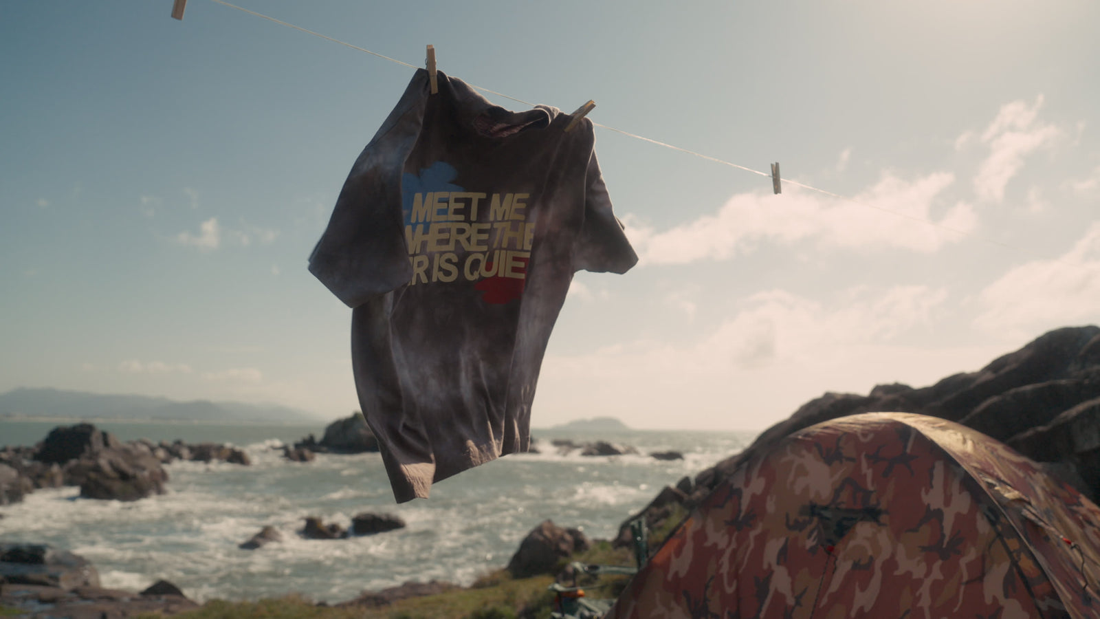 Camiseta “Meet me where the air is quiet” pendurada no varal, com o mar e a barraca ao fundo